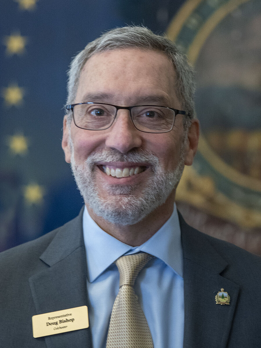 Smiling man in glasses wearing a suit with a nameplate, against a blue and gold backdrop.