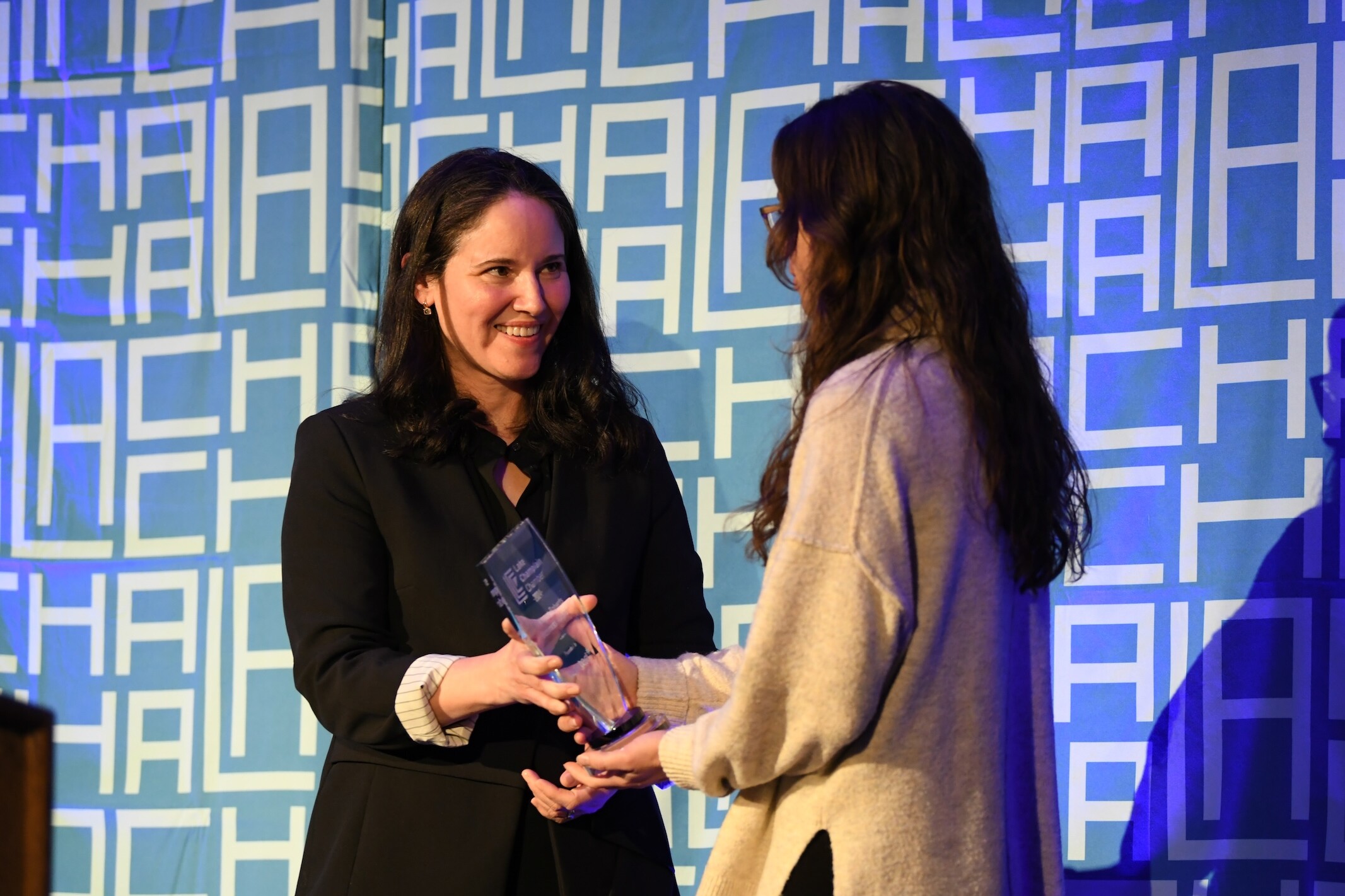 Two women are smiling as one hands the other an award on stage.