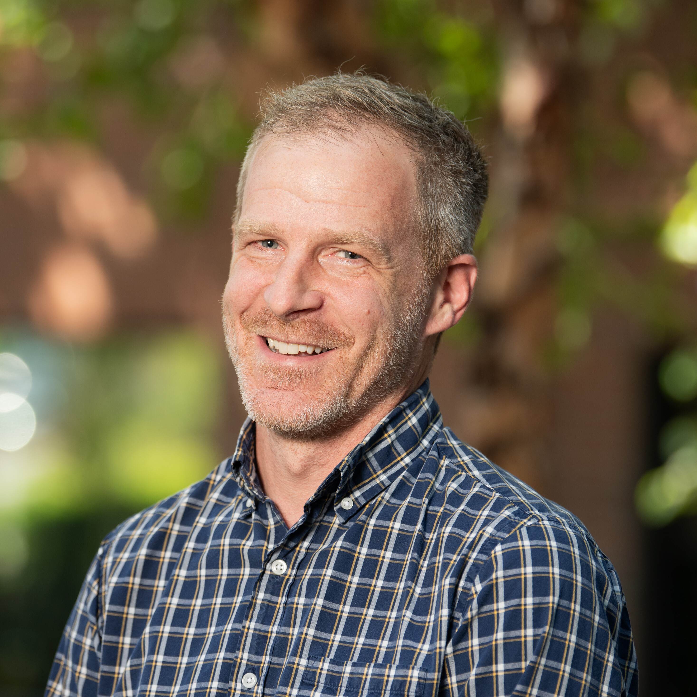 Smiling man in a plaid shirt stands outdoors with greenery in the background.