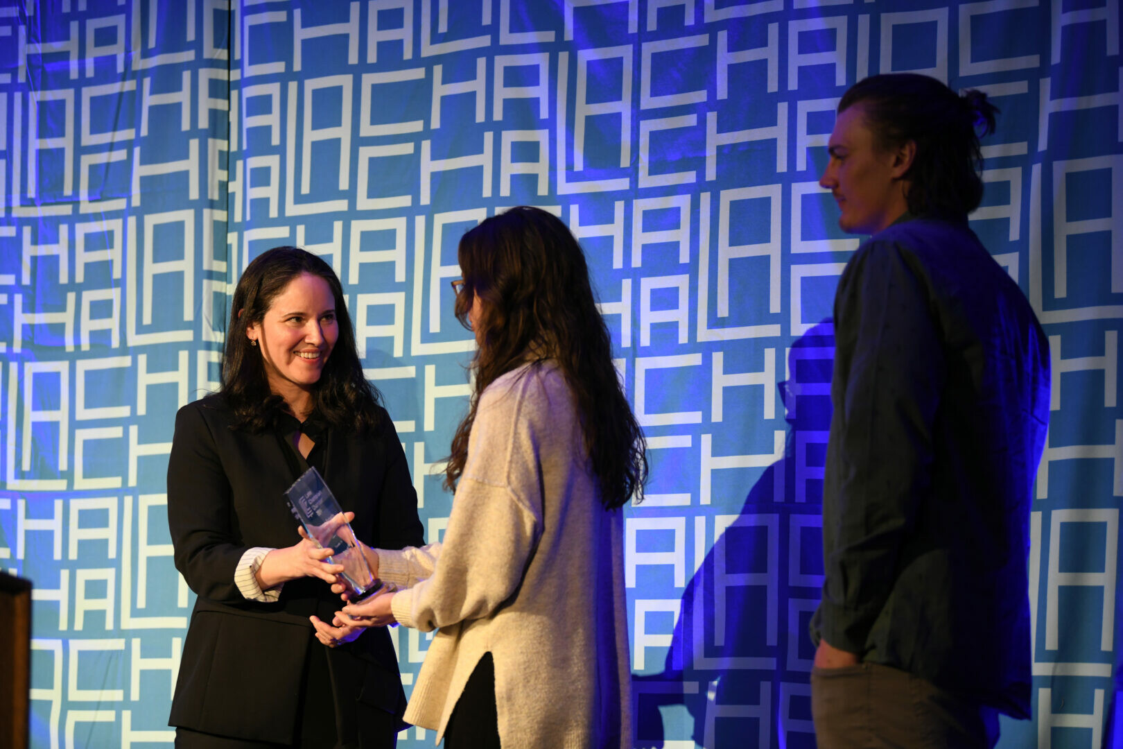 A woman hands an award to another woman on stage, with a man watching.
