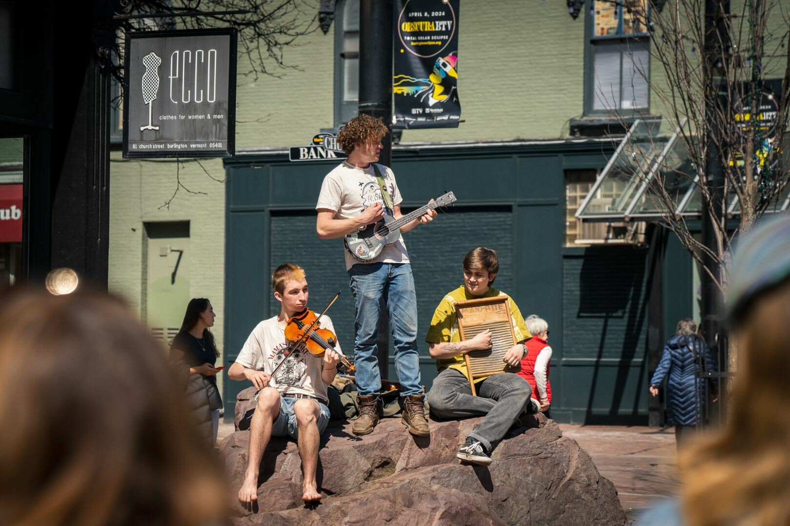 Three musicians perform on a rock, playing guitar, violin, and washboard outdoors.