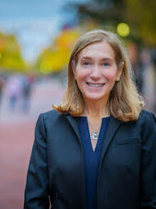 Smiling woman in a dark blazer, outdoors with blurred pedestrians in the background.