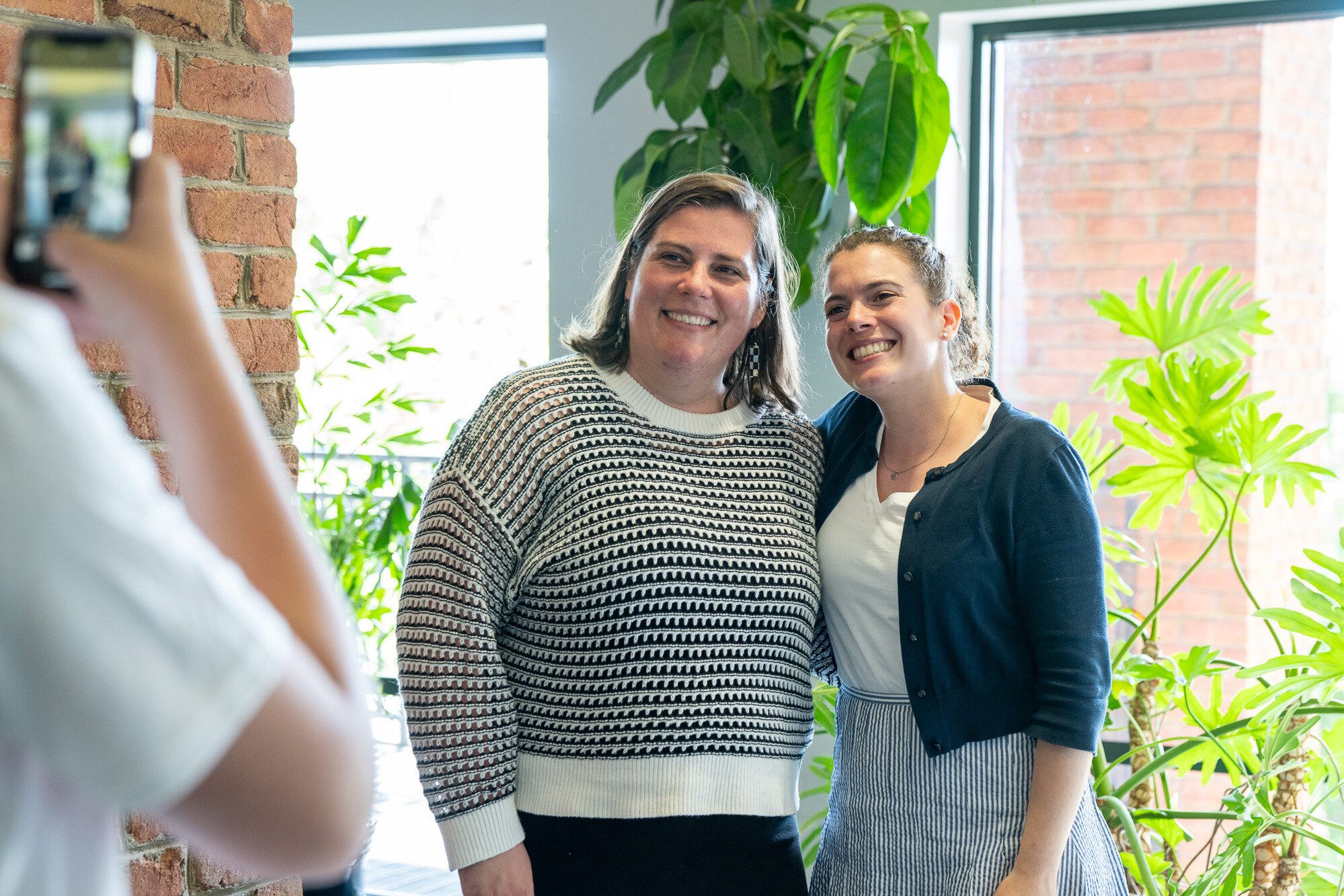 Two women smile together for a photo in a bright, plant-filled indoor space.