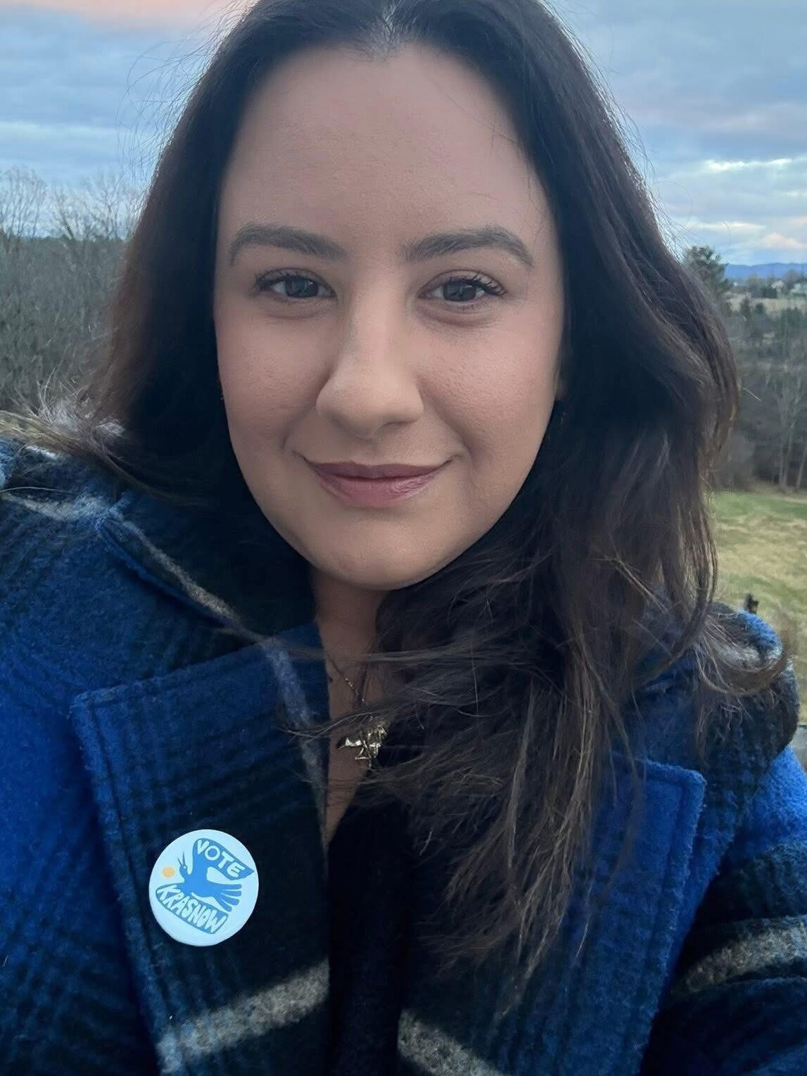 A woman in a blue coat smiles, wearing a "Vote" badge outdoors.