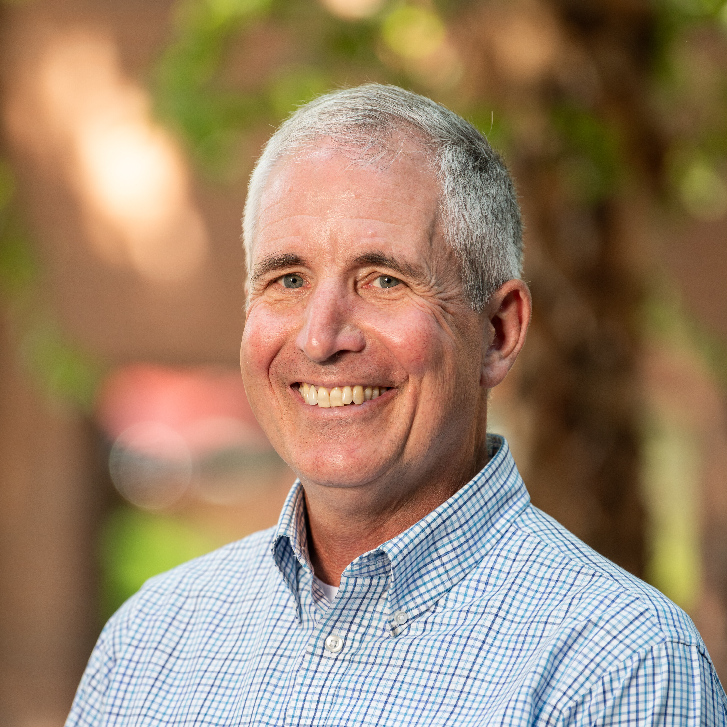 Smiling man in a checkered shirt outdoors with greenery in the background.