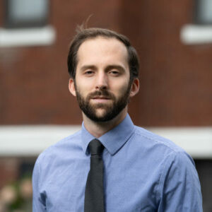 A man with a beard stands outdoors, wearing a blue shirt and a tie.