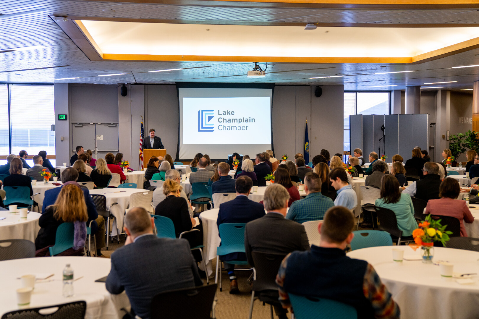 A speaker presenting at a Lake Champlain Chamber event with attendees seated at tables.