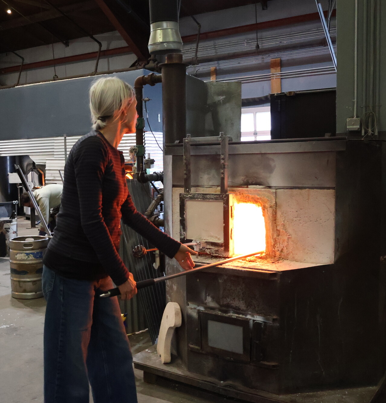 A woman works with a furnace, using a rod to manipulate glowing material inside.