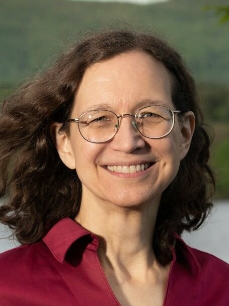 Smiling woman with glasses, wearing a red shirt, outdoors by a body of water.