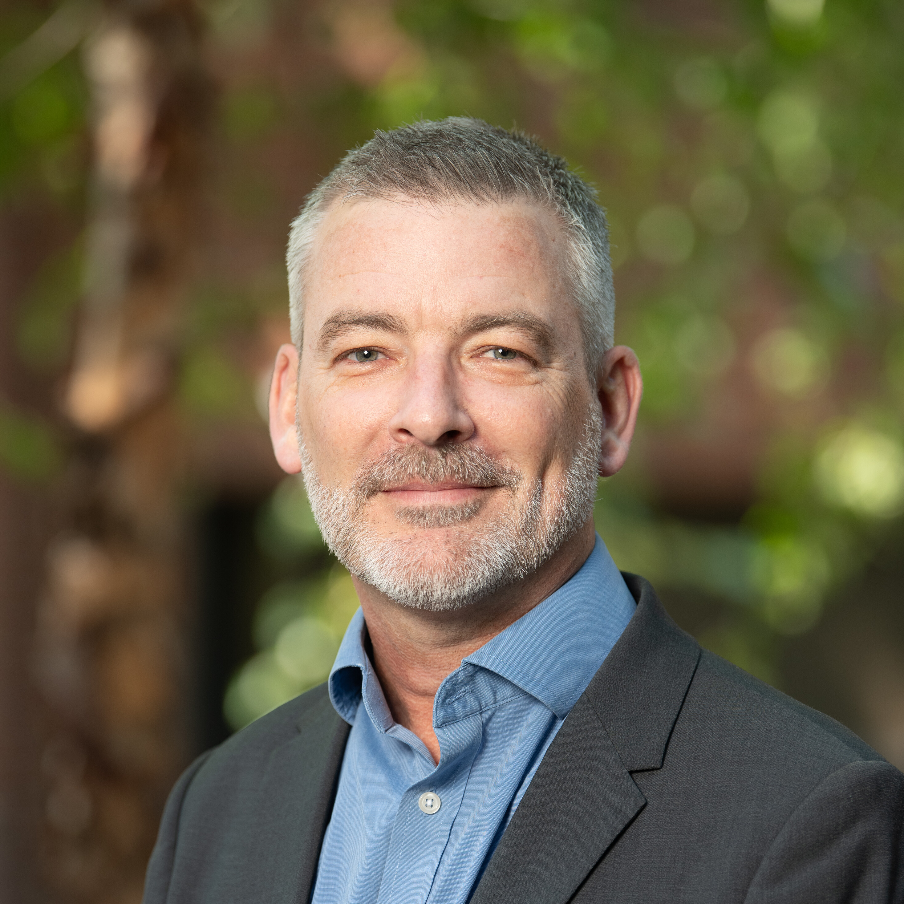 Man with gray hair and beard in a suit, smiling outdoors with greenery in the background.