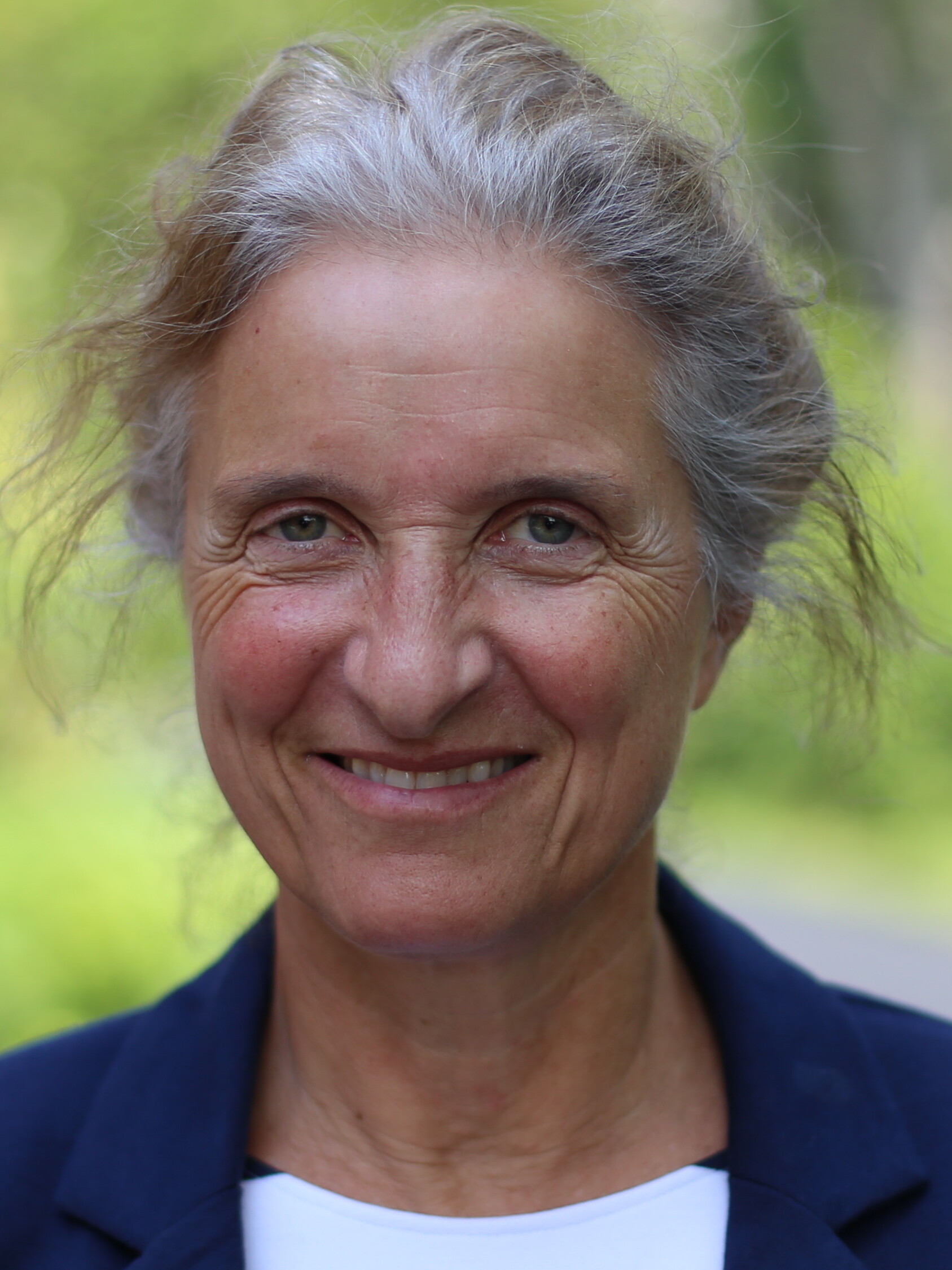 Smiling middle-aged woman with gray hair, outdoors, wearing a navy blazer.