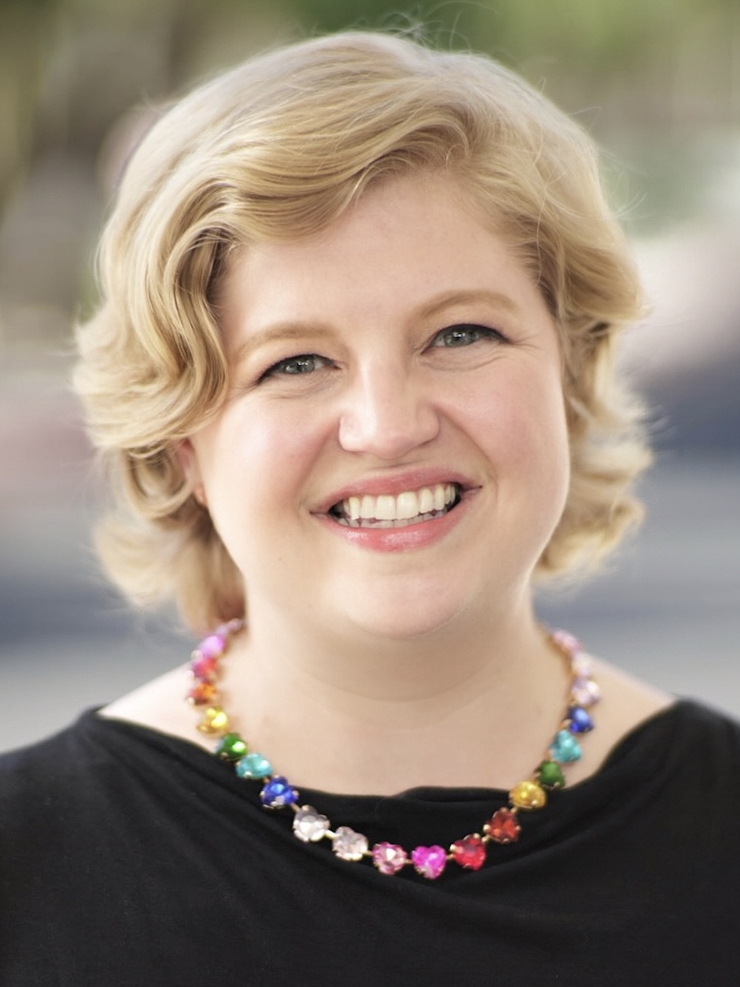 Woman with short, wavy hair smiles brightly, wearing a colorful beaded necklace.