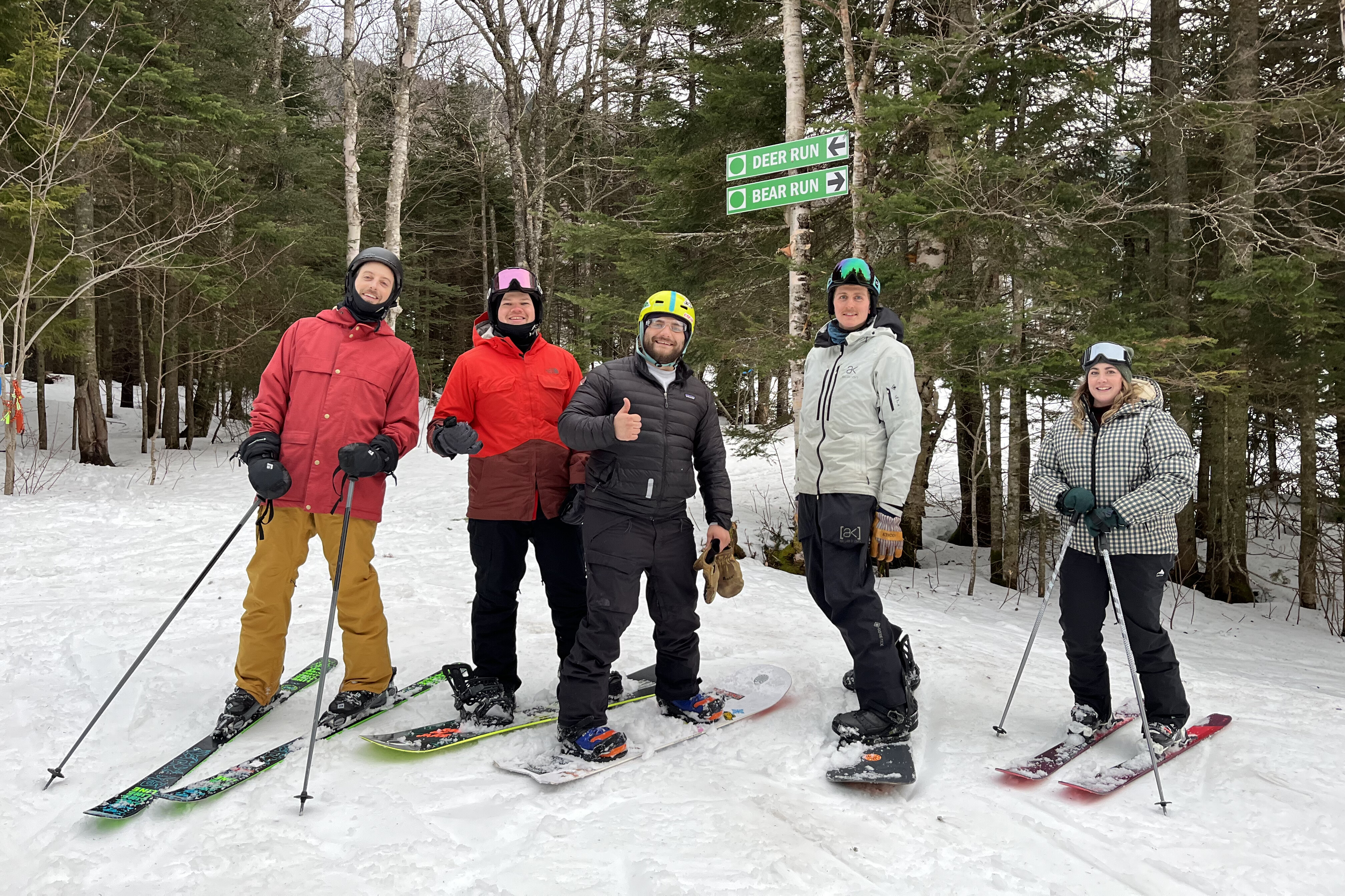 Group of five skiers posing with snow-covered trees and trail signs in the background.