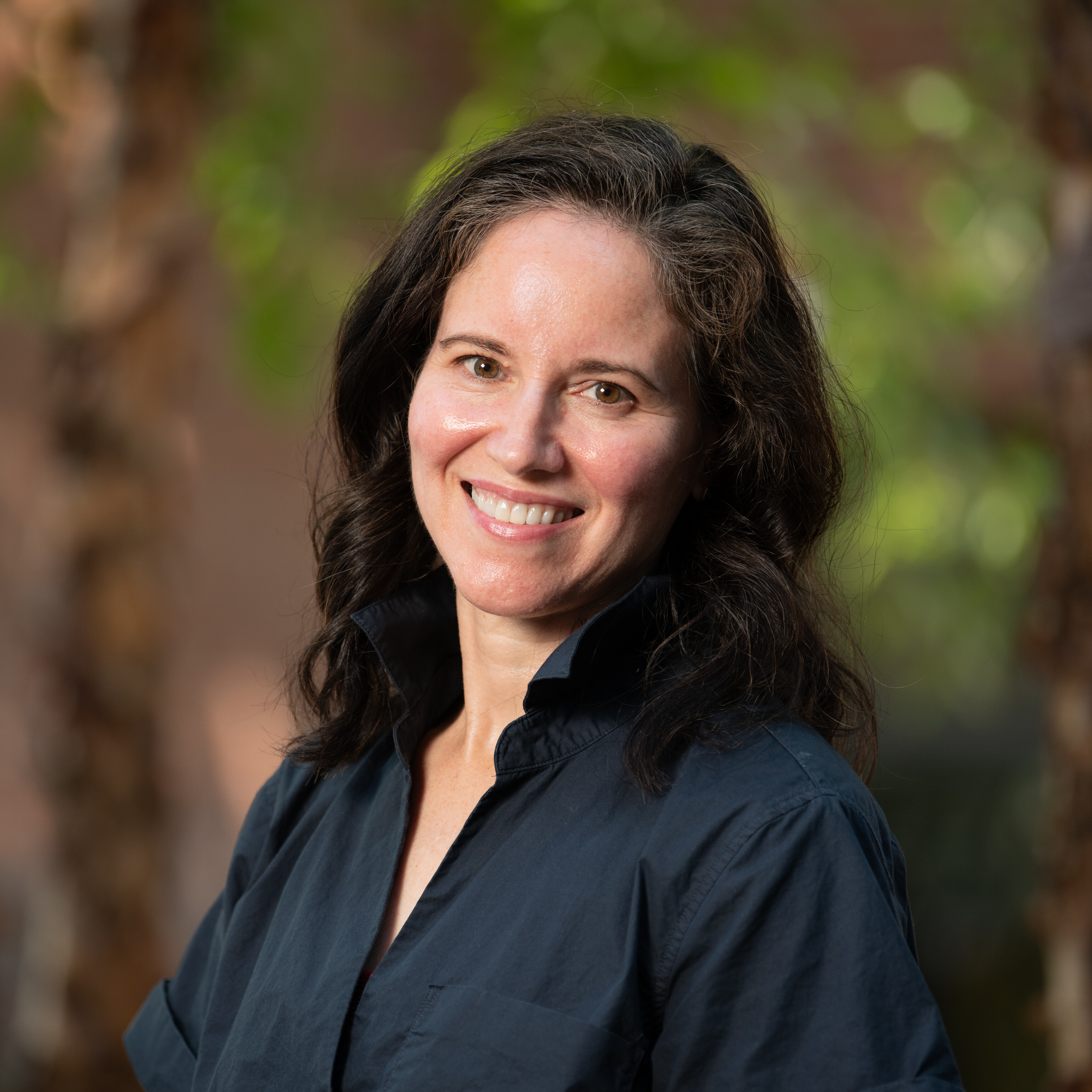 Woman with dark hair smiles, wearing a black shirt, with greenery in the background.