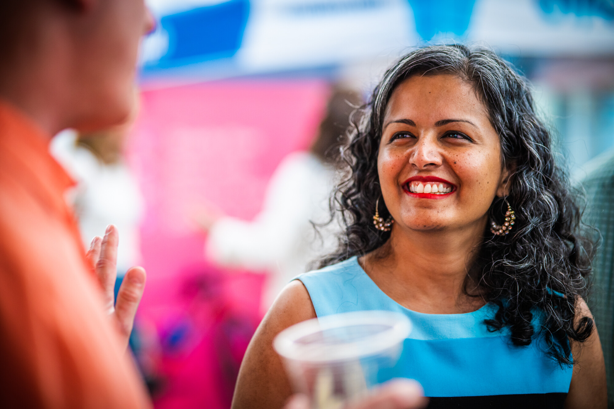 A smiling woman in a blue dress engages in conversation at an outdoor event.