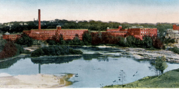 Historic red brick factory building beside a calm river and green landscape.