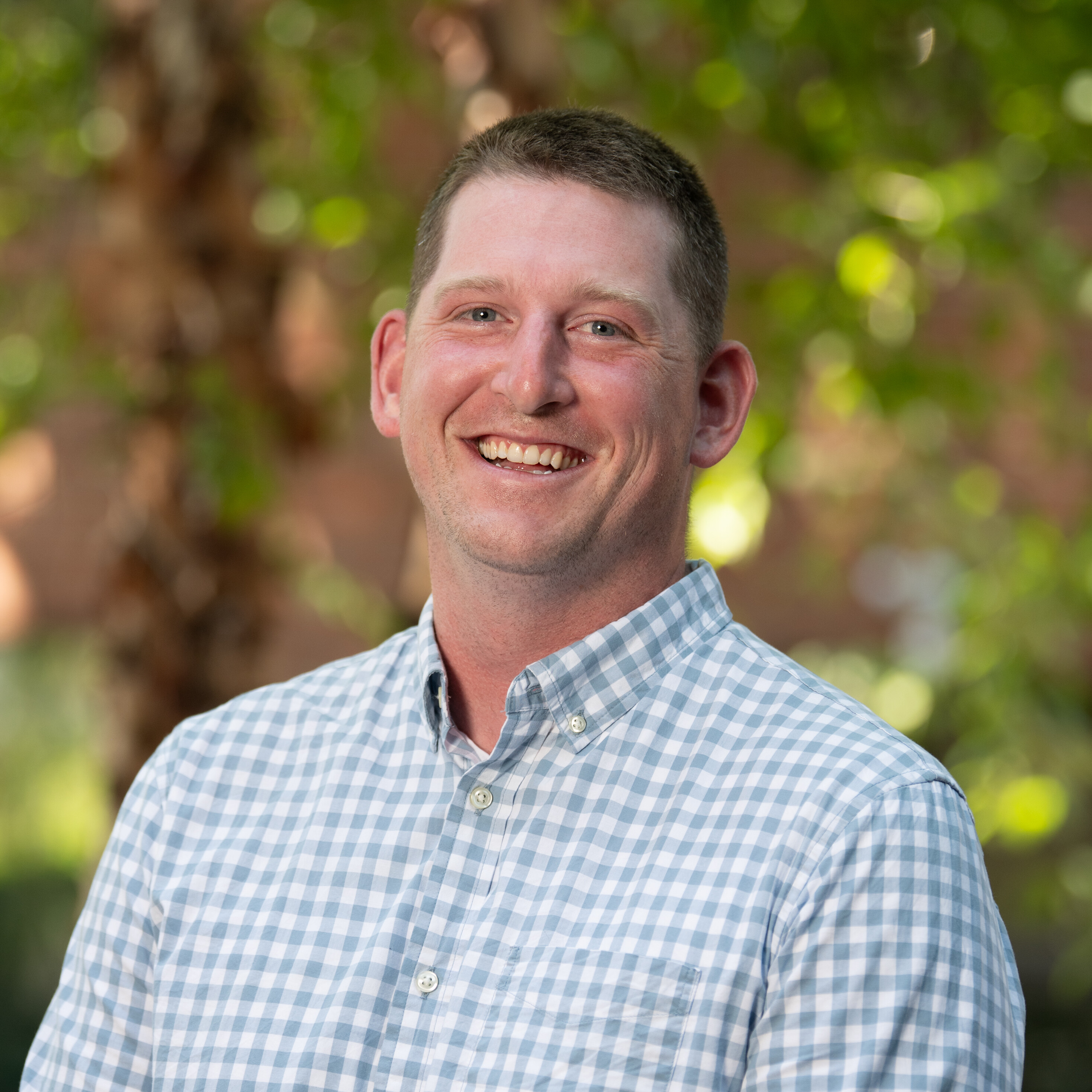 Smiling man in a checkered shirt, with greenery in the background.