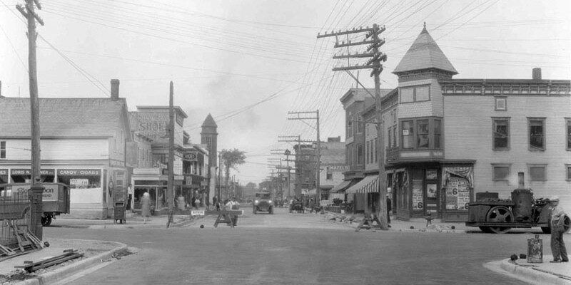 Historic black-and-white street scene featuring shops and early 20th-century vehicles.