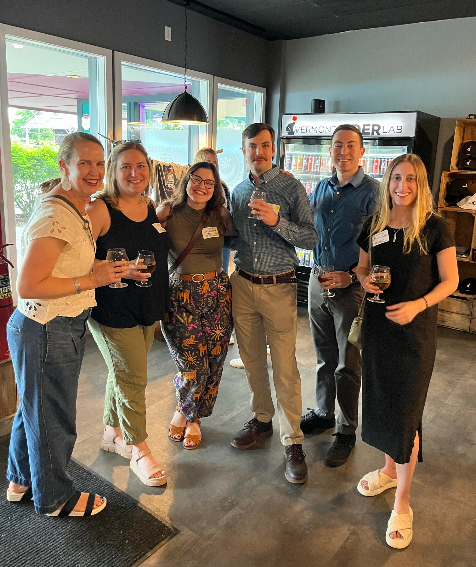 Group of six people holding drinks, smiling, in a casual indoor setting.