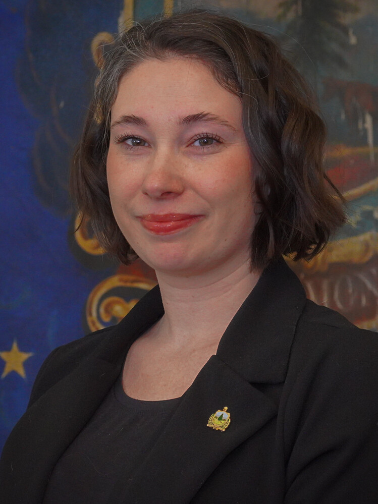 A woman with short curly hair wearing a black blazer poses in front of a colorful background.