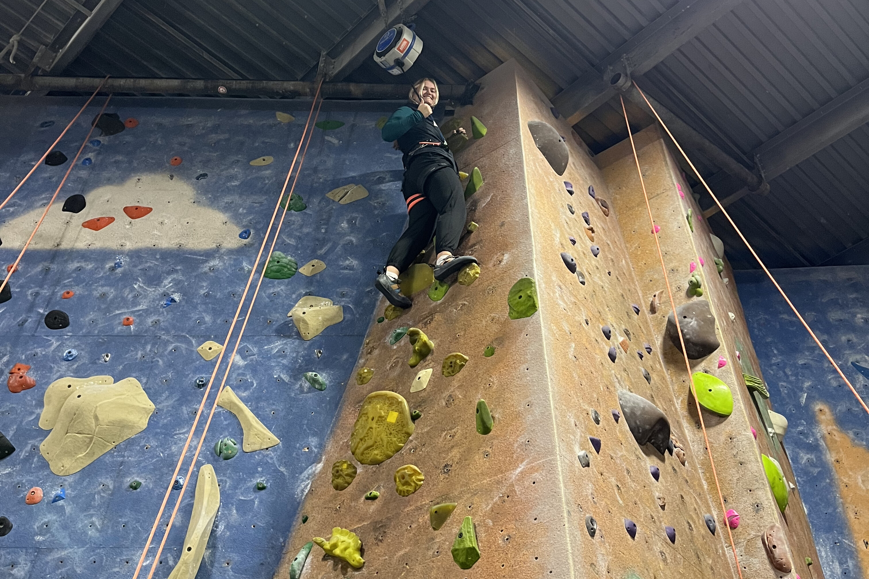 Person climbing an indoor rock wall, smiling with climbing gear and colorful holds.