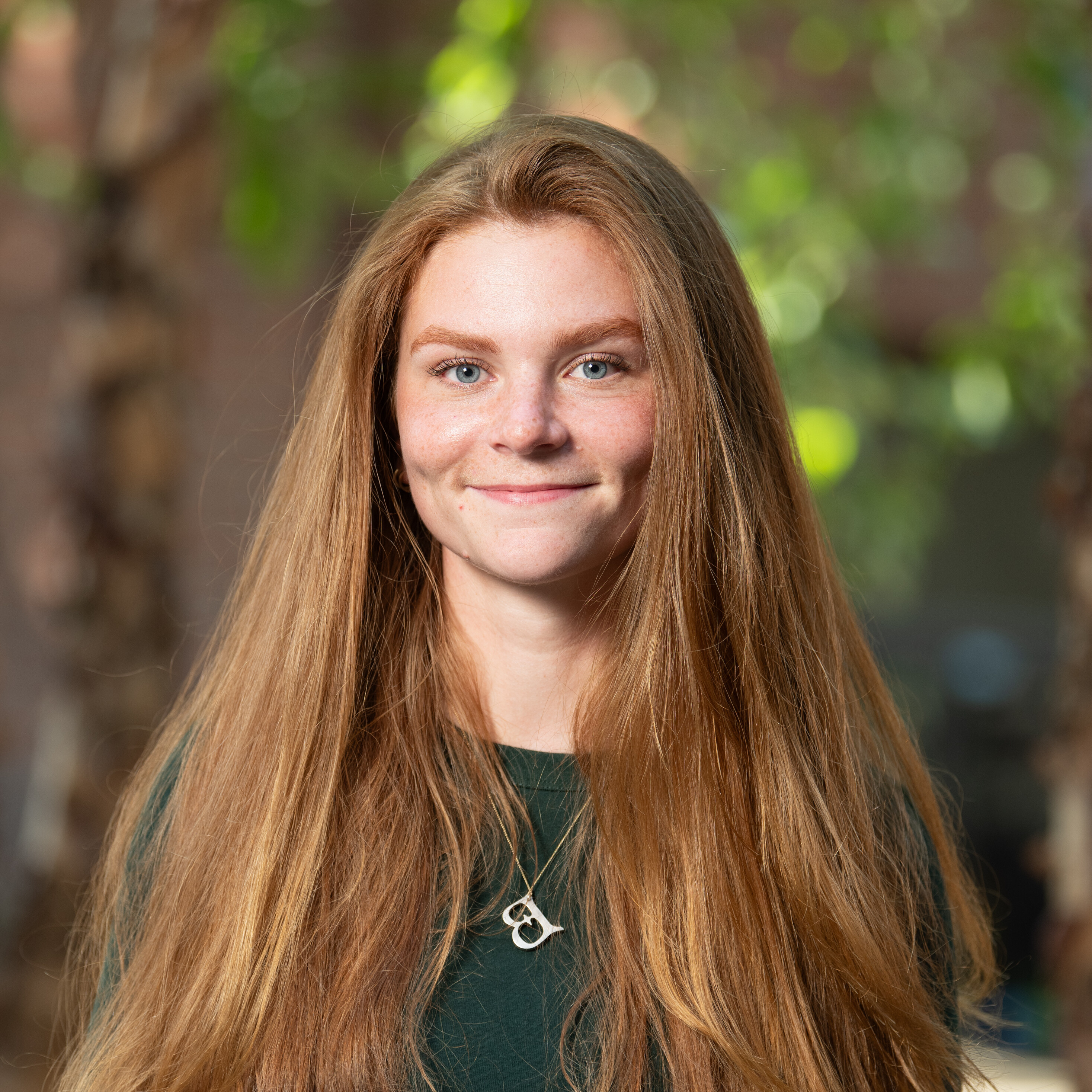 A smiling young woman with long, straight hair poses outdoors, wearing a green shirt and necklace.