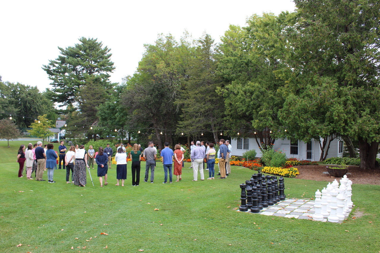 Group of people gathered outdoors near a large chess set and colorful flower beds.