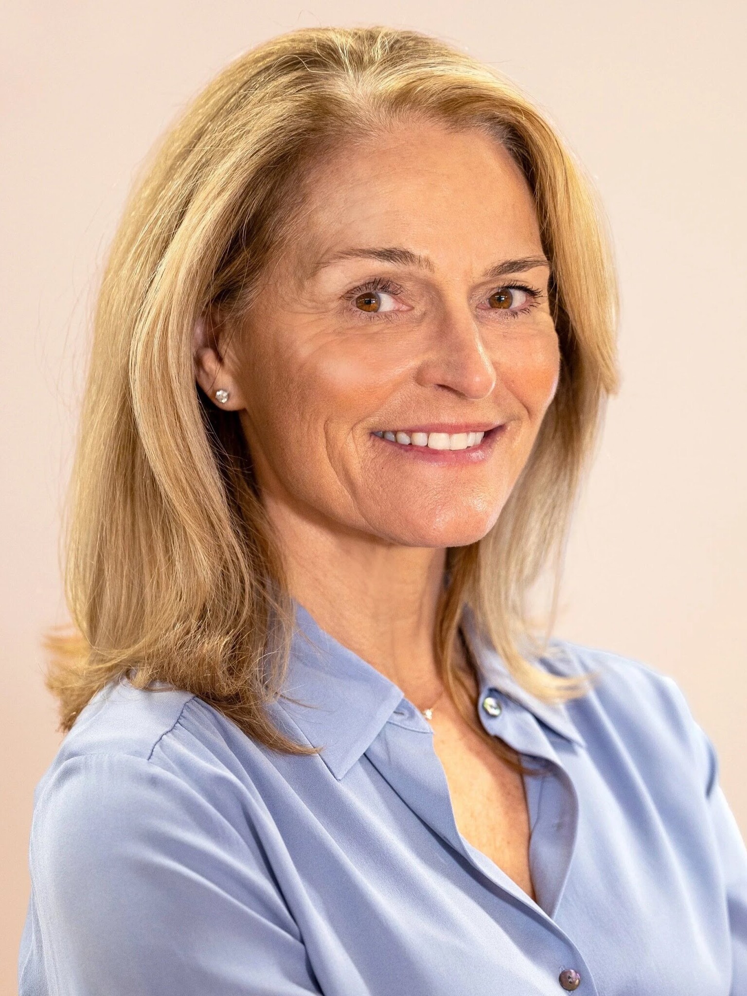 Smiling woman with long blonde hair, wearing a light blue shirt against a soft background.