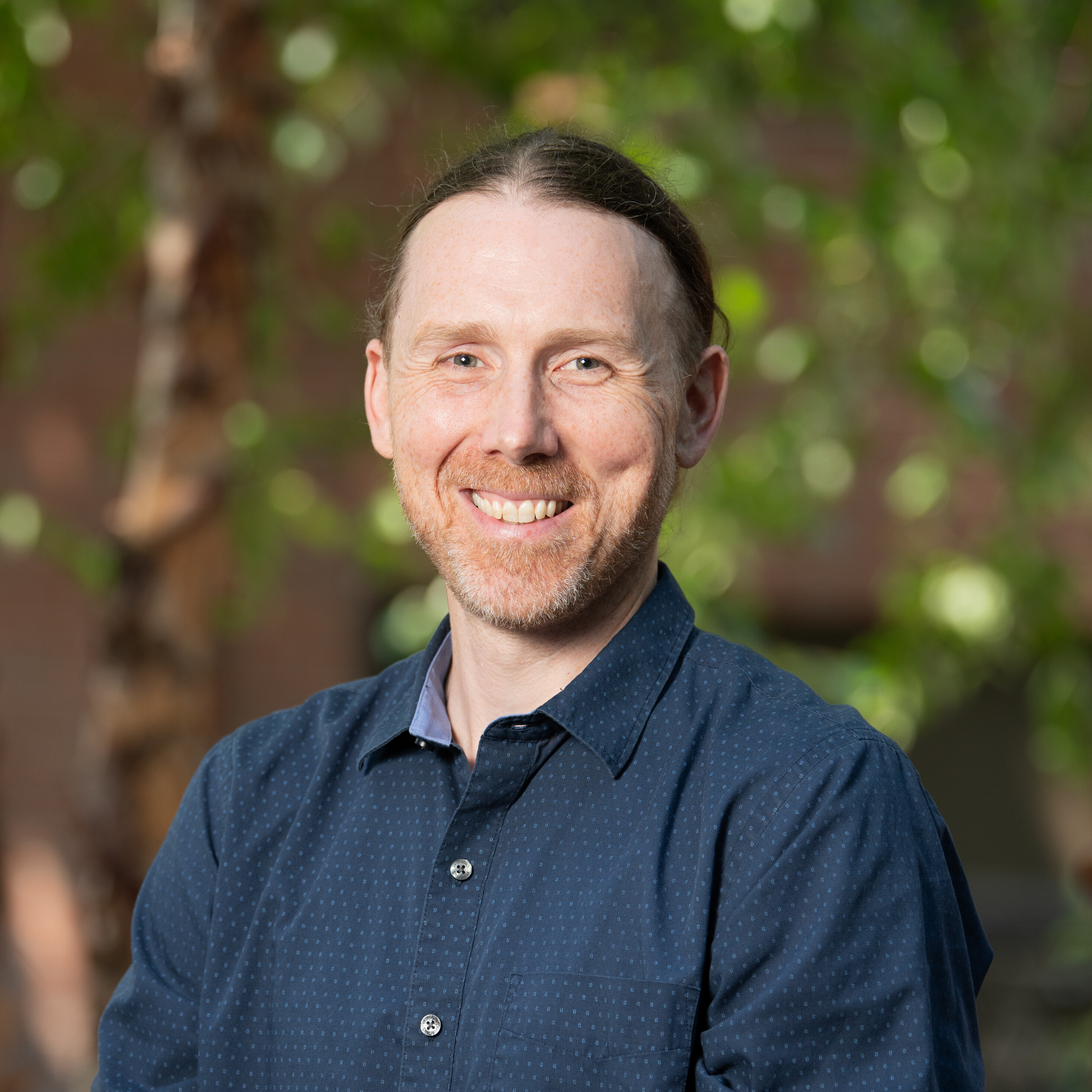 Smiling person in a blue shirt, outdoors with green foliage in the background.