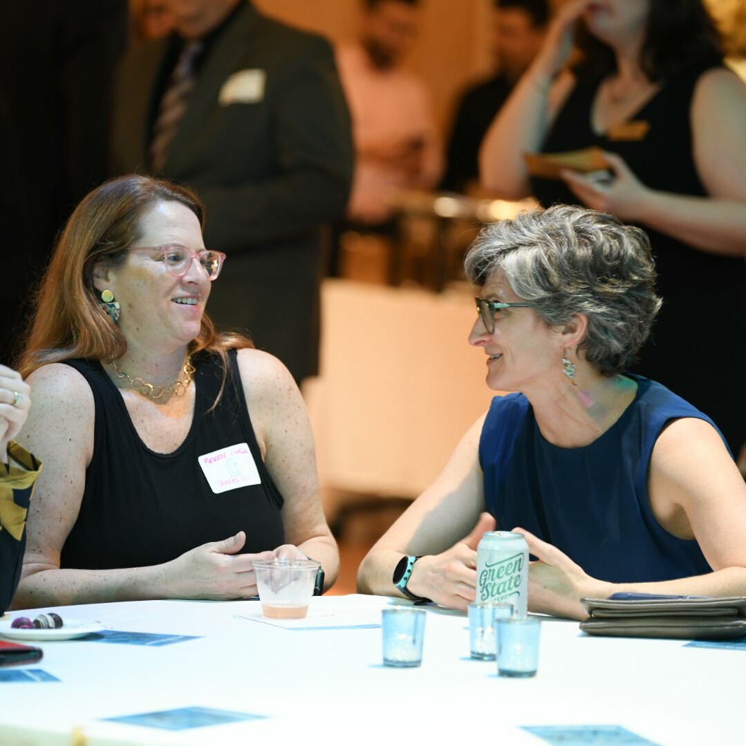 Two women chat at a table, enjoying drinks in a lively event setting.