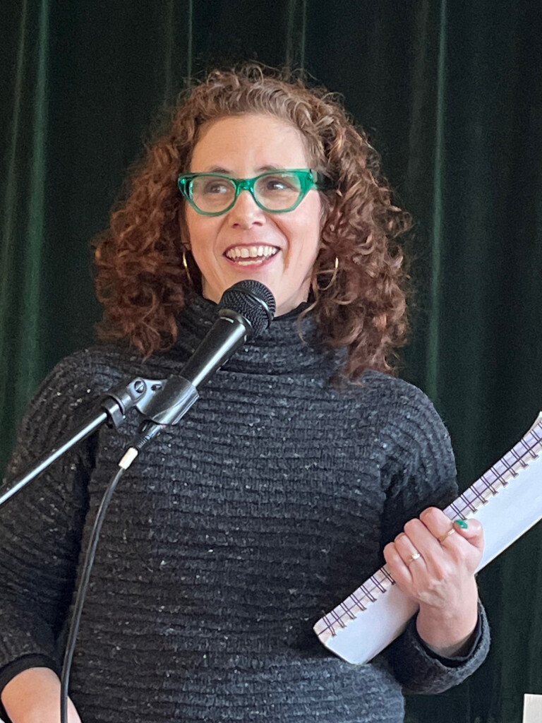 Woman with curly hair and green glasses speaks into a microphone, holding notes.