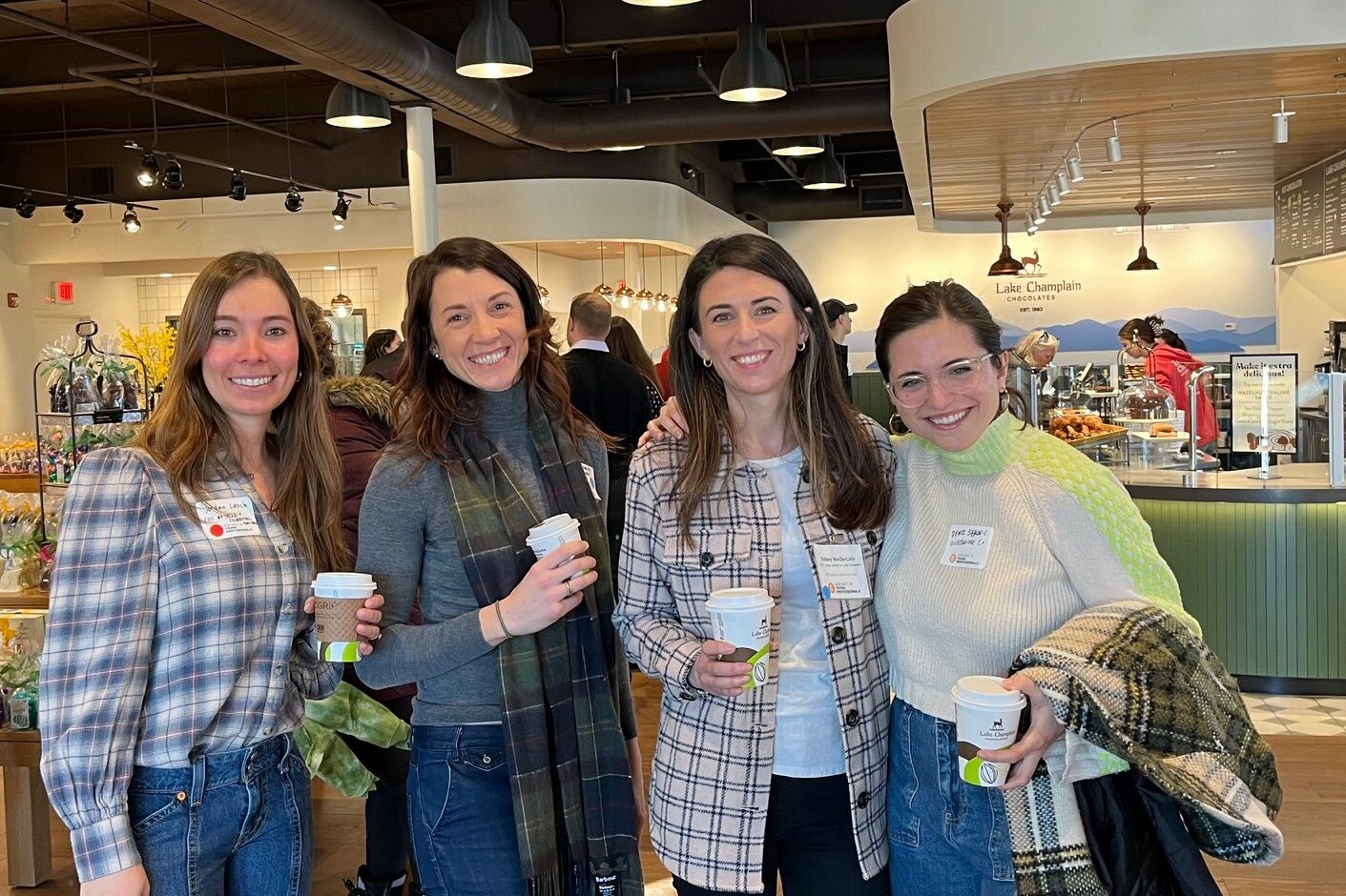 Four women smiling in a caf&eacute;, each holding coffee cups, with a modern decor backdrop.