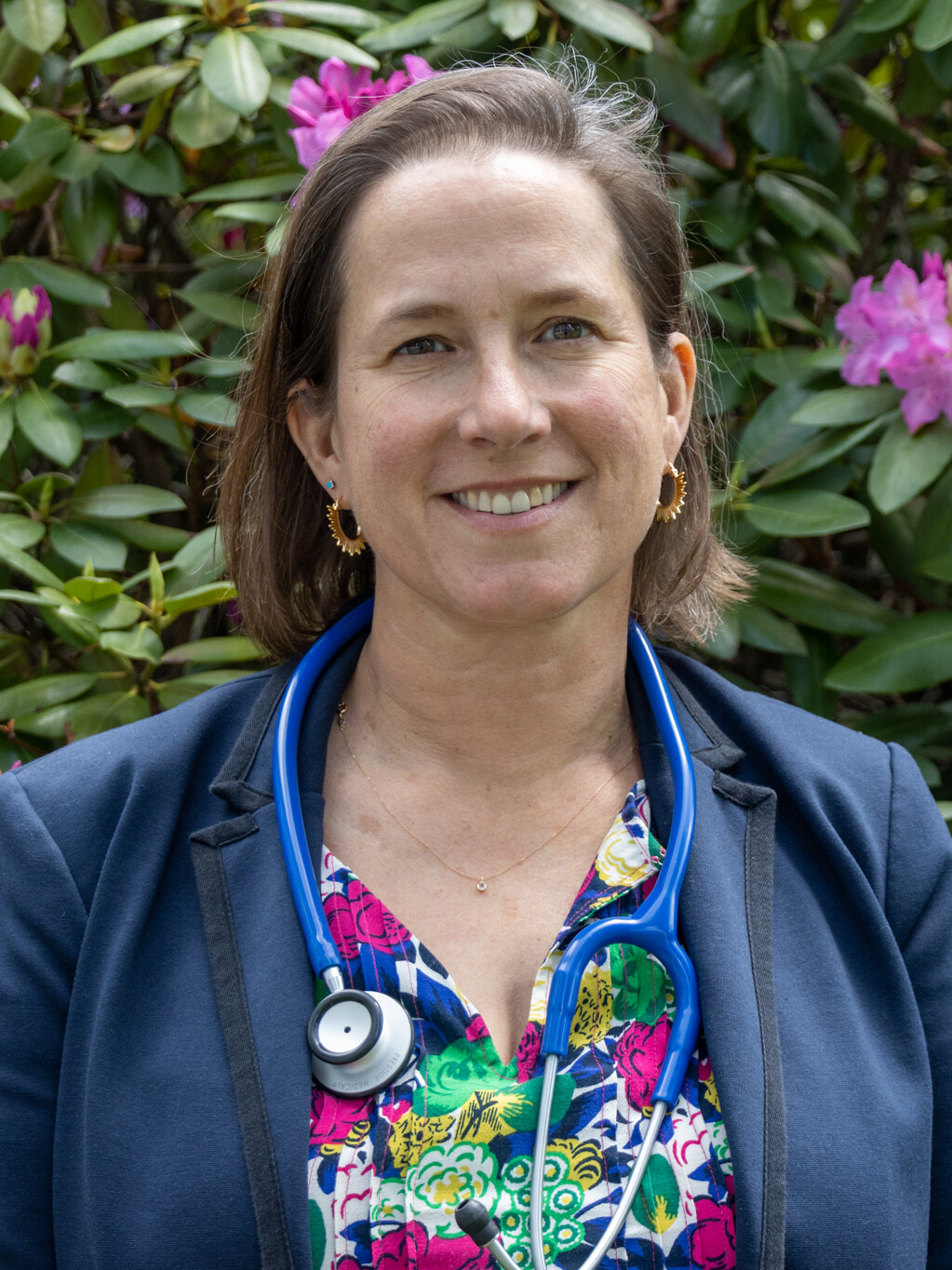 Woman with a stethoscope smiling in front of flowering greenery.