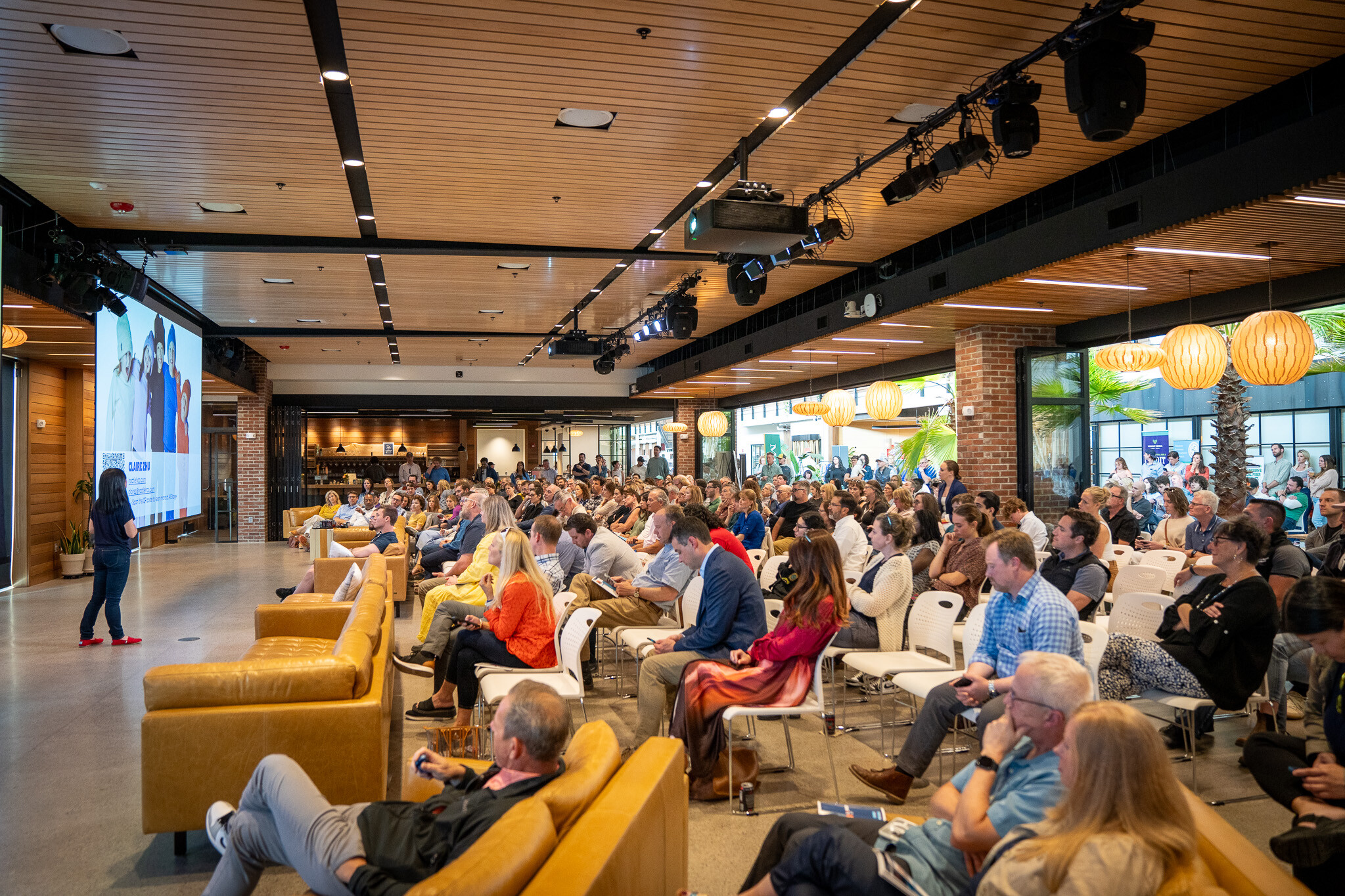 A speaker presents to a large audience in a modern indoor venue with seating.