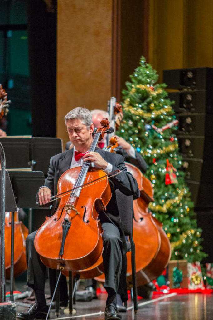 photo of cellist playing wearing a red bowtie with a christmas tree behind him