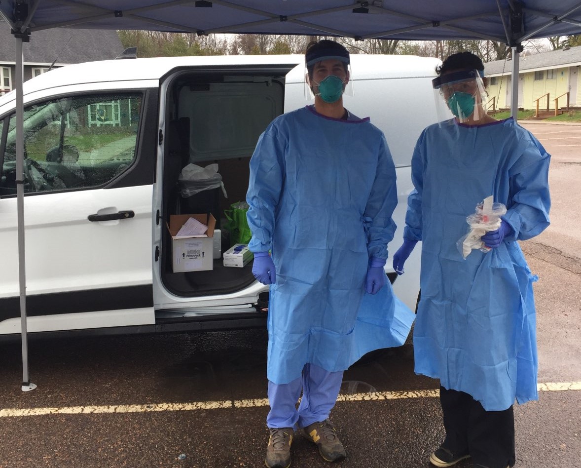 two healthcare workers in full personal protective equipment stand in front of a van